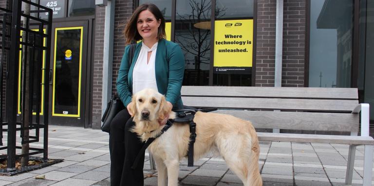 Une femme souriante assise sur un banc avec son chien-guide d'INCA, à l'extérieur d'un édifice d'INCA avec des affiches et des affiches jaunes d'INCA.