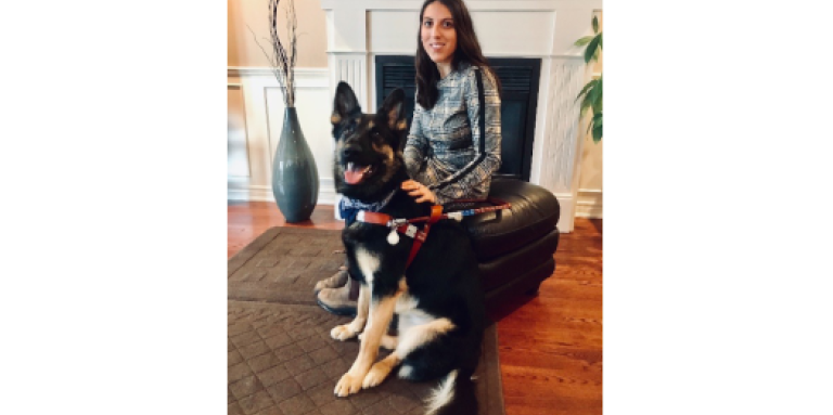 Stela sitting on a leather ottoman in front of a fireplace with her German Shepherd guide dog in front of her.