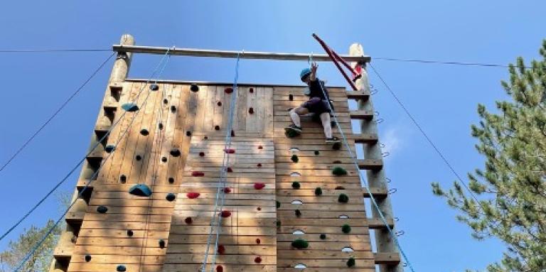 Julia scales the climbing tower at CNIB Lake Joe. She is wearing a harness and helmet. 