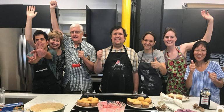 Sept personnes lèvent les bras dans les airs fièrement, derrière un comptoir de cuisine rempli de cupcakes, de glaçage, d'ingrédients et d'ustensiles de cuisine. Un homme tient un canne blanche.