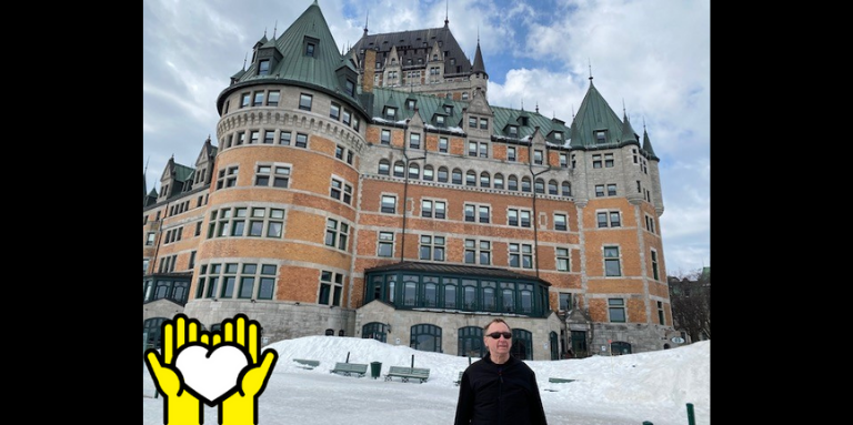 Une photo de Louis Lévesque debout en face du Château Frontenac à Québec. L'icône de deux mains tenant un cœur blanc sur un fond jaune, dans le coin inférieur gauche.