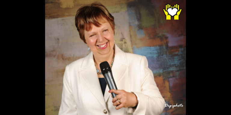 Une photo de Louise Burley souriant et tenant un micro de sa main gauche.  L'icône de deux mains tenant un cœur blanc sur un fond jaune, dans le coin supérieur droit.