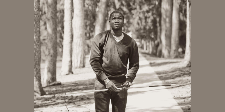 A black and white photograph of Ben walking in a forest. He holds his white cane up in the air near his waist.