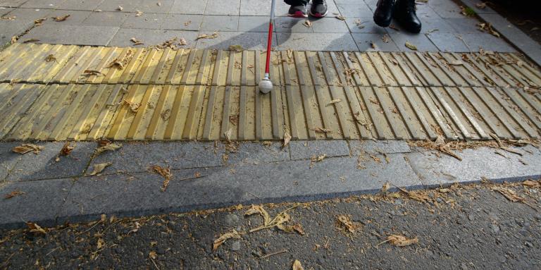 Une canne blanche avec un embout rond tournant balaie le pavée tactile d'un trottoir.
