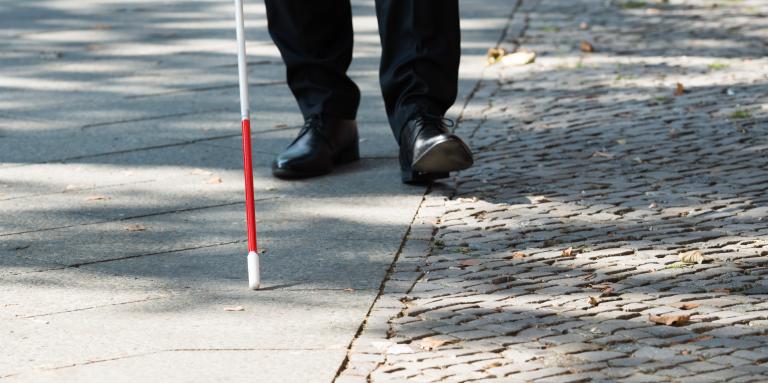 Une canne blanche balaie un trottoir. En arrière-plan, des chaussures d'homme suivent le bout de la canne