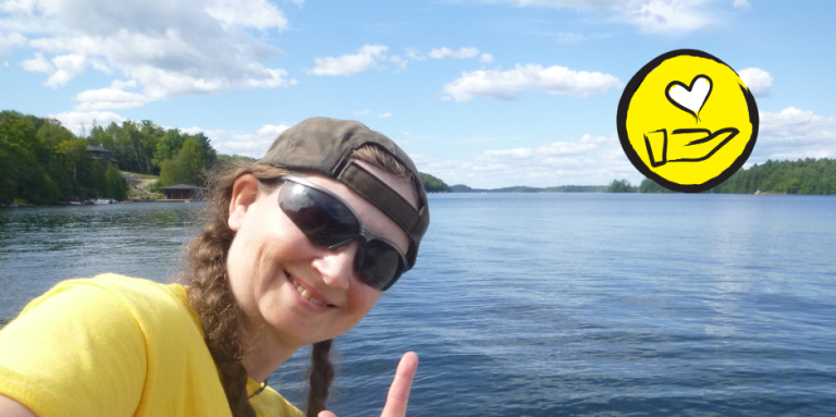 Jessica Bailey pose en face du lac Joe. Elle fait le signe en langue des signes américaine pour "Je t'aime". L'icône d'une main surmontée d'un cœur flottant apparaît dans le coin supérieur droit. 