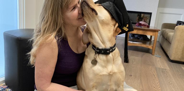 Laurie est assise sur le sol de son salon avec son chien-guide, Bridget, un croisement de Labrador Retriever jaune et de Golden Retriever. Bridget porte un chapeau de graduation et est assise sur les genoux de Laurie. Laurie presse affectueusement son visage contre le visage duveteux de Bridget.