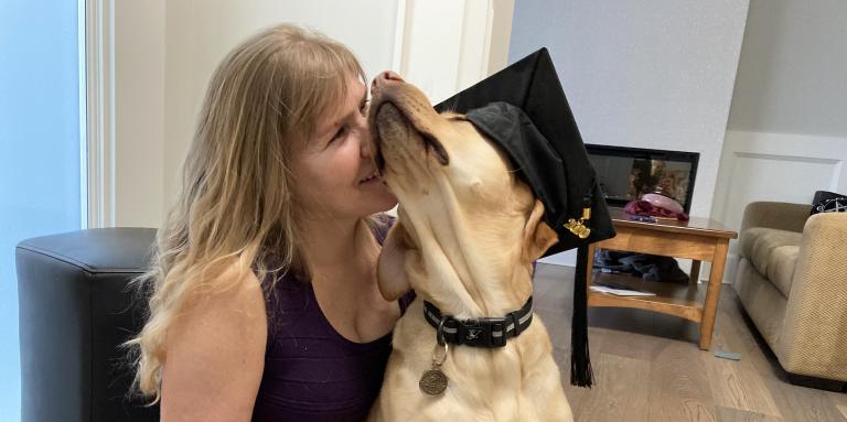 Laurie est assise sur le sol de son salon avec son chien-guide, Bridget, un croisement de Labrador Retriever jaune et de Golden Retriever. Bridget porte un chapeau de graduation et est assise sur les genoux de Laurie. Laurie presse affectueusement son visage contre le visage duveteux de Bridget.