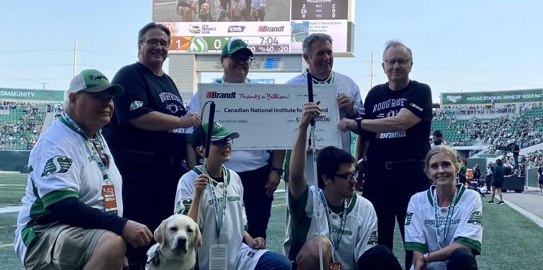 Des représentants d'INCA et du groupe d’entreprises Brandt posent ensemble pour une photo alors qu'ils tiennent le chèque de 2 millions de dollars lors du match des Roughriders de la Saskatchewan. Deux chiens-guides d'INCA portent des chandails verts