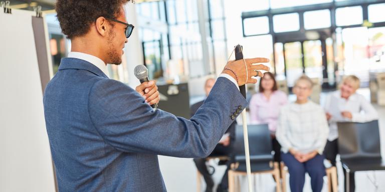 A young man stands at the front of a meeting room and speaks to a group of people. He is holding a microphone in his left hand and a white cane in his right hand.