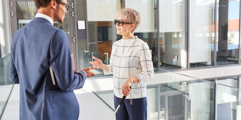 Deux employés discutent dans un bureau lumineux. L'une des employées, une femme d'âge moyen aux cheveux courts et gris, utilise une canne blanche.