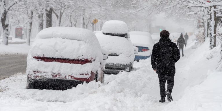 Une rue résidentielle, des trottoirs et des voitures garées sont recouverts de neige. Un homme marche sur un trottoir enneigé.  