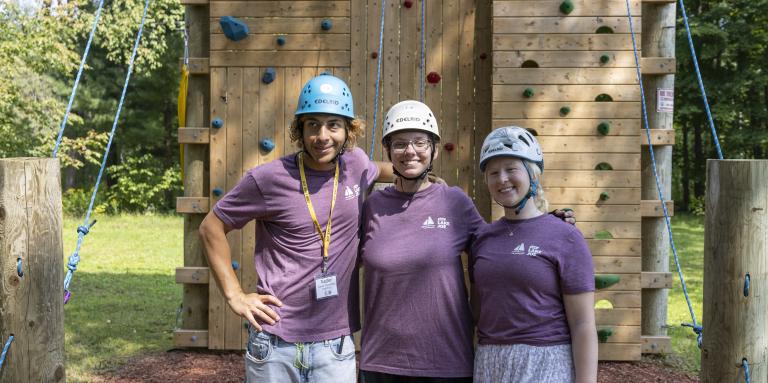 Three staff members stand in front of the accessible climbing tower. 