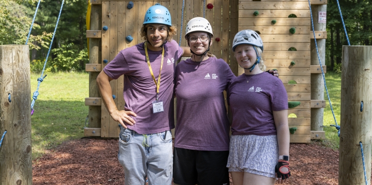 Three staff members stand in front of the accessible climbing tower. 