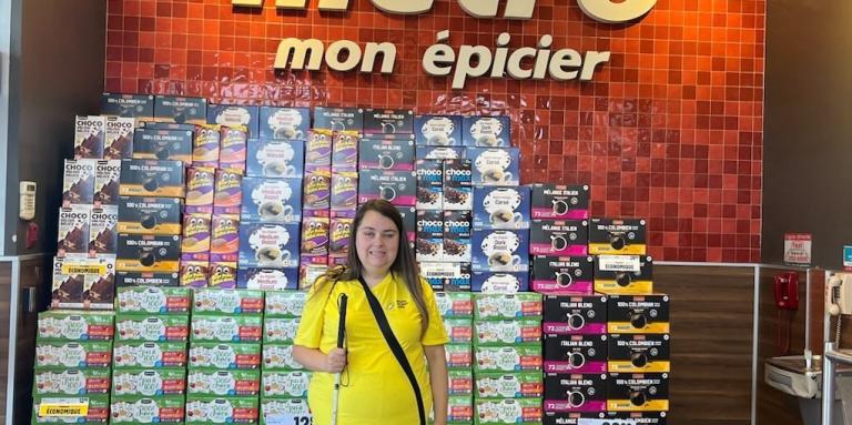 Carolane portant son chandail Quartier Accessible devant l'insigne de l'épicerie Métro avec à la main droite sa canne blanche