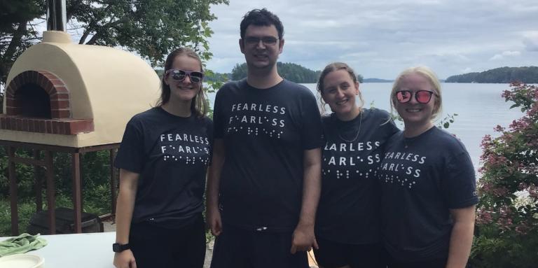 De gauche à droite : (G-D) Emilee Schevers, Tavish Gillan, Taylor Gaudon et Jade Ondrik près du four à pizza extérieur au Camp Lake Joe d'INCA. Ils portent des t-shirts bleus sur lesquels figure le mot « Fearless » en lettres moulées et en braille.