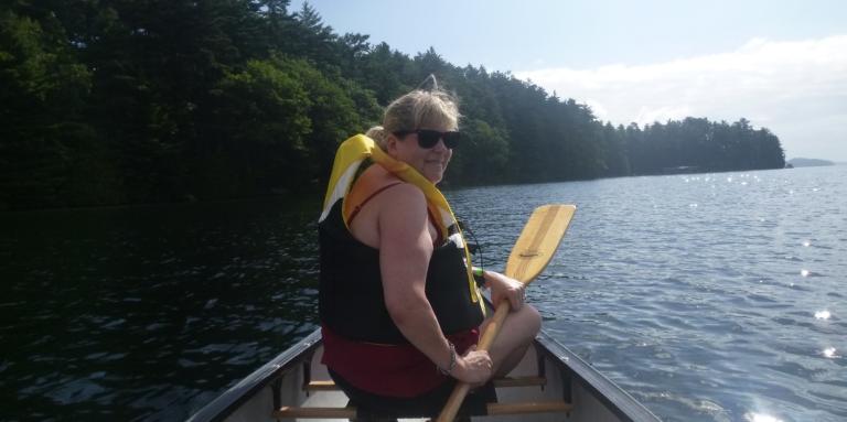 Khrisstina sitting in the front of a canoe holding a paddle in her hand. She is turning around to smile at the camera. The water is sparkling and there are green trees on her left.