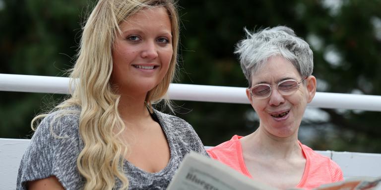 Une aînée aveugle accompagnée d'une jeune femme, assises sur un banc dans un parc, qui lisent le journal