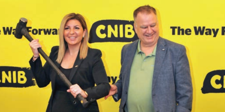 Angela Bonfanti, Présidente et chef de la direction d'INCA, et Robert J. Fenton KC, président du conseil d'administration, debout ensemble devant l'iconographie d'INCA. Angela tient un marteau qui signifie briser les barrières – toutes deux sourient.