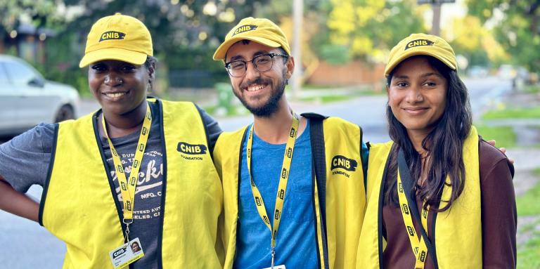 Trois collecteurs de fonds travaillant pour le compte d'INCA sourient à la caméra, debout sur le trottoir dans un quartier résidentiel. Chaque collecteur de fonds porte une casquette de baseball jaune d'INCA, un gilet et un cordon avec photo d'identité.