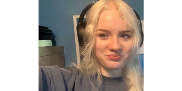 Acacia, une jeune femme aux cheveux blancs, sourit et pose pour une photo de type selfie. Elle porte des écouteurs sur les oreilles et un long sweat-shirt gris.