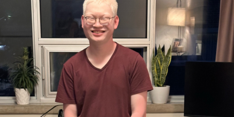 Aidan, un jeune homme aux cheveux blancs, se tient devant un bureau à domicile/un poste de travail informatique. Aidan porte des lunettes et un t-shirt marron avec un jean. Il sourit et se tient debout, les mains jointes.