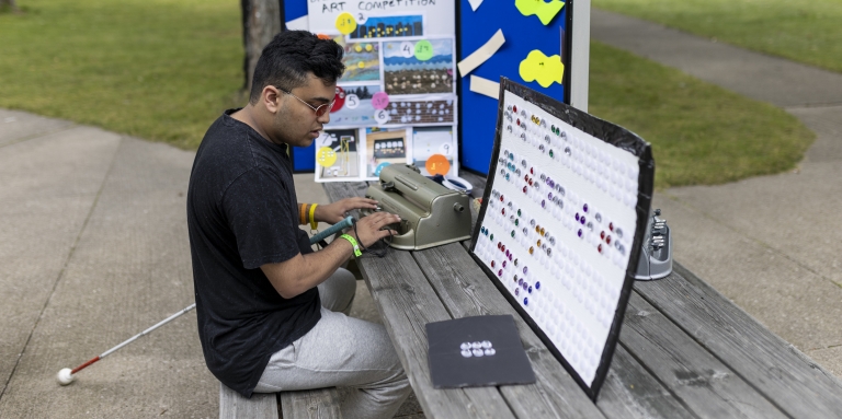 Un jeune homme est assis à une table de pique-nique au Centre Lake Joe et tape sur une machine braille Perkins. Il porte des lunettes de soleil et sa canne blanche repose sur le banc de la table de pique-nique. À sa gauche se trouve un grand panneau de présentation bleu avec des informations sur le braille.