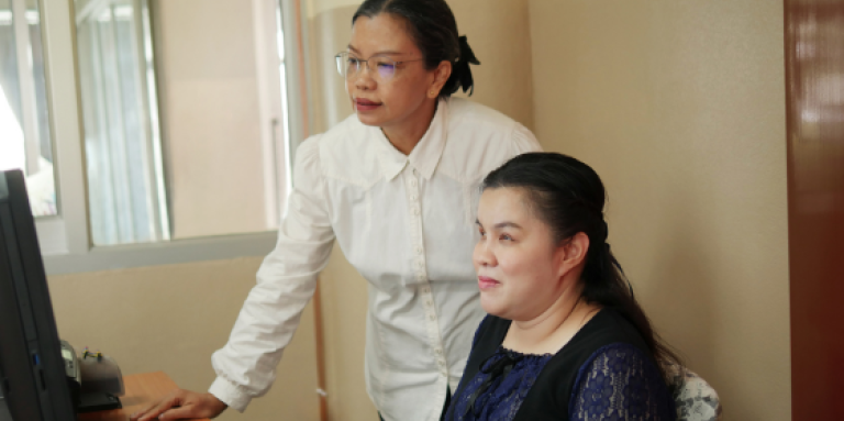 Dans un bureau, une femme est assise à un pupitre et utilise une afficheur braille rafraîchissable reliée à un ordinateur. Une collègue se tient à sa droite, le bras appuyé sur le bureau. Ils passent en revue un document professionnel sur l'écran de l'ordinateur.