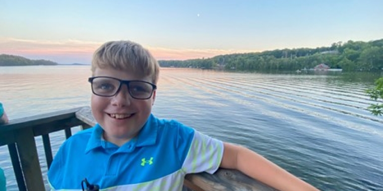 Un jeune garçon portant des lunettes et tenant une canne blanche s'appuie contre une balustrade en bois. Derrière lui, un lac entouré d’arbres à l'horizon.