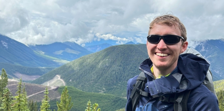 Un jeune homme se tient en plein air, sur une colline et on aperçoit des montagnes en arrière-plan. Il porte des lunettes de soleil noires, une veste bleue, un sac à dos et une canne est attachée à sa ceinture.