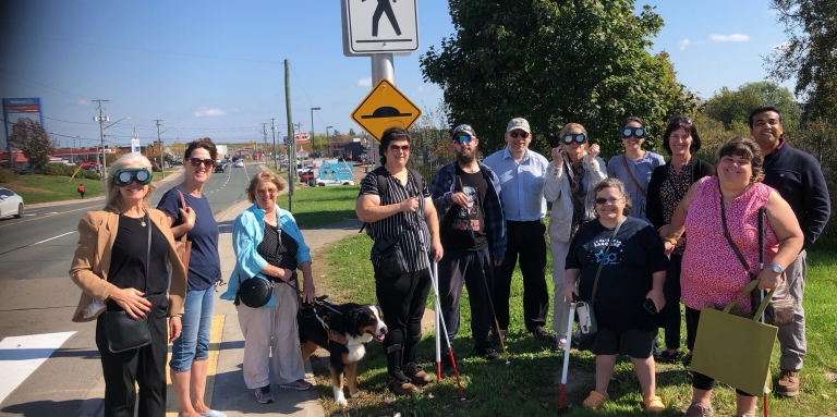 Un grand groupe de personnes souriantes se tient à côté d'un passage pour piétons situé près d'une grande route L'une des personnes a un chien d'aveugle, plusieurs autres portent des lunettes qui simulent différentes expériences de la cécité.