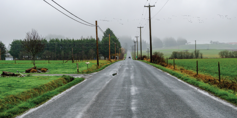 Une longue route rurale droite qui s'étend au loin. C'est un jour de brouillard et il y a des flaques d'eau sur la chaussée. De chaque côté de la route, il y a des champs, des arbres et des lignes électriques. 
