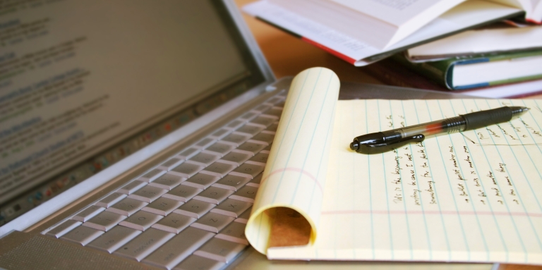 Vue d'un bloc-notes jaune avec écriture, un ordinateur portable ouvert et un assortiment de livres sur le bureau