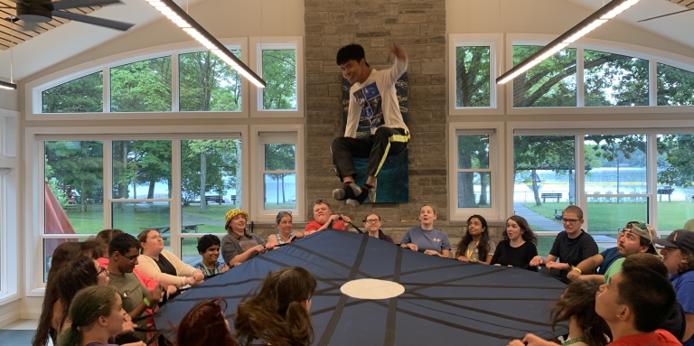 Des campeurs d'été tiennent un trampoline en cercle après avoir lancé un campeur dans les airs.