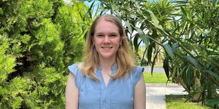 Laura, une jeune femme aux cheveux blonds, sourit devant un jardin verdoyant avec des palmiers et des arbustes. Elle porte une robe de soleil bleu clair.
