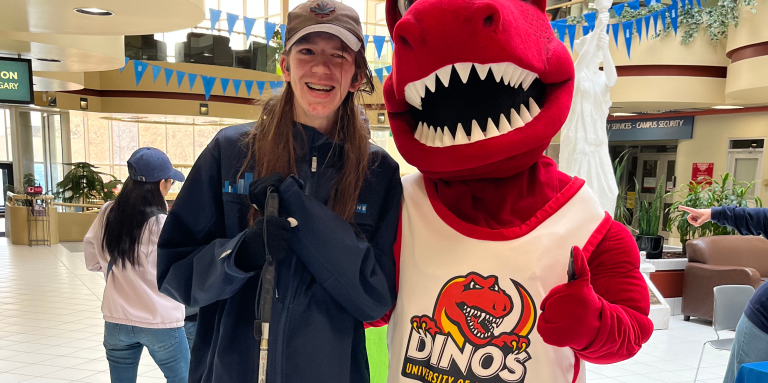 Zachary, un jeune homme aux longs cheveux bruns, sourit à côté de Rex, la mascotte dinosaure rouge et duveteuse de l'université de Calgary. Zach est vêtu d'un sweat-shirt bleu, d'un pantalon de survêtement, d'un chapeau et tient sa canne blanche.