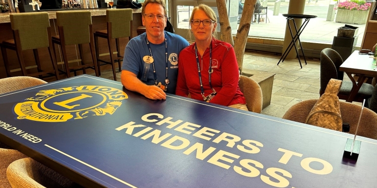 Jerry et Jenn sont assis ensemble derrière une table sur laquelle sont imprimés le logo du Lions International et les mots "Cheers to Kindness".