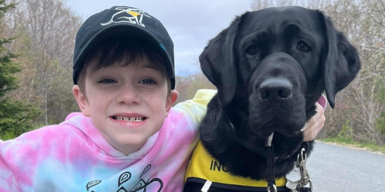 Un jeune garçon à la peau claire sourit à la caméra tout en s'accroupissant sur une route à côté d'un chien noir qui porte un dossard de La Course à pattes. Le garçon tient une canne blanche dans sa main droite et son bras gauche entoure le chien.
