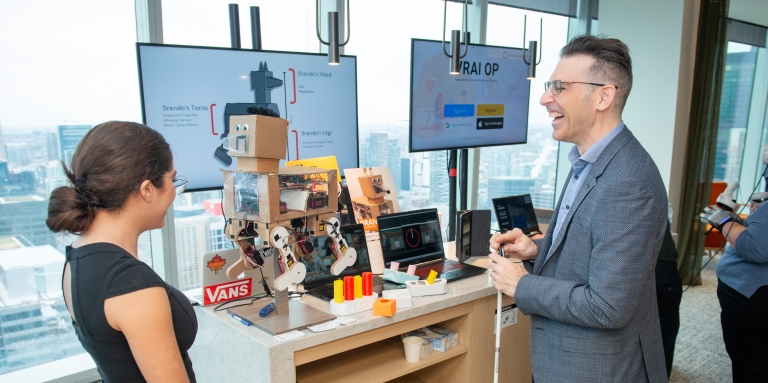 Un homme en costume tenant une boîte blanche s'adresse à une femme devant une table remplie de technologies robotiques.