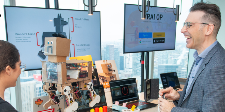 Un homme en costume tenant une boîte blanche s'adresse à une femme devant une table remplie de technologies robotiques.