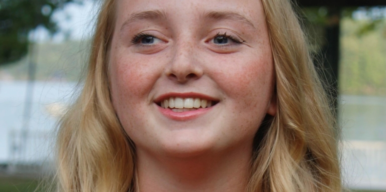 Portrait de Maggie, souriante, pris en extérieur à Lake Joe, près du front de mer. Maggie est une jeune femme aux longs cheveux blonds qui porte une chemise orange.