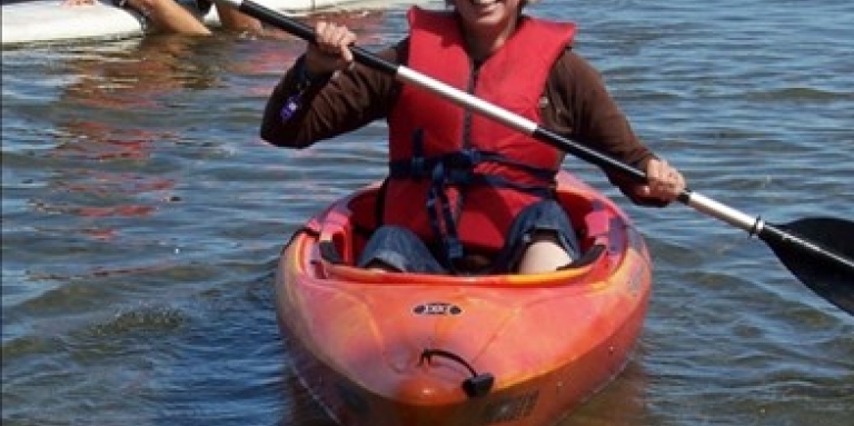 Joanne sourit et pose dans un kayak orange pour une photo sur le lac. Elle porte des lunettes de soleil, un gilet de sauvetage rouge et tient sa pagaie.