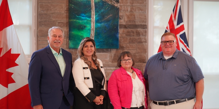 L'honorable Neil Lumsden, ministre des Sports de l'Ontario, Angela Bonfanti, présidente-directrice générale de l'INCA, Sharon Cadeau, vice-présidente de la Nation métisse de l'Ontario, et l'honorable Graydon Smith, député provincial de Parry Sound–Muskoka, se tiennent devant la cheminée du centre Lake Joe de l'INCA. Les drapeaux du Canada et de l'Ontario sont à nos côtés.