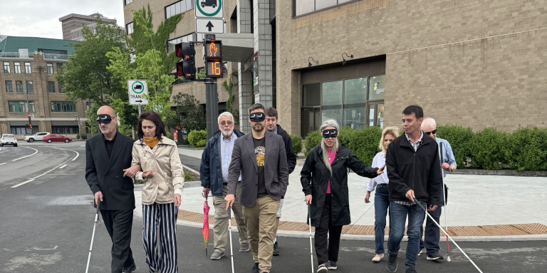 un groupe de personnes qui traversent la rue, y'en a qui ont les yeux bandés qui ont une canne et qui sont guidés par soit une personne ayant une limitation visuelle soit une personne voyante.
