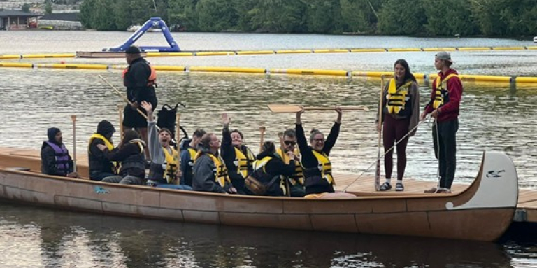 Un groupe de personnes dans un grand canoë Voyageur à quai. Elles portent des gilets de sauvetage jaunes et ont les mains levées en l'air.