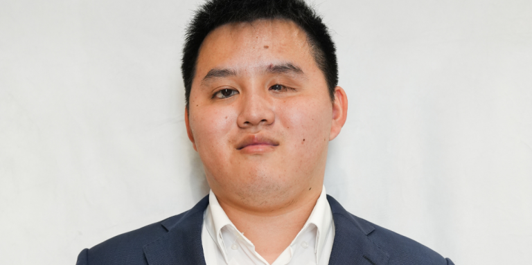 Zhicheng, un jeune homme asiatique vêtu d'un costume bleu marine et d'une chemise blanche, se tient debout devant un fond blanc. Zhicheng sourit et la photographie ressemble à un portrait professionnel.