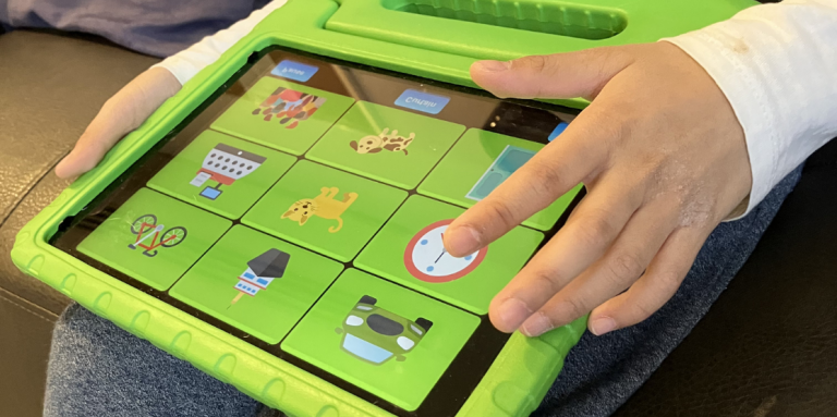 Les mains d'un enfant tiennent un iPad dans un étui de protection vert vif et tapotent une grille d'icônes colorées représentant notamment des animaux, des objets et une horloge à l'écran.
