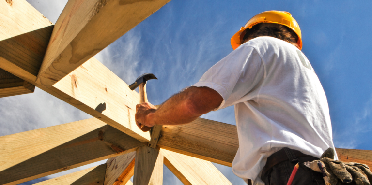 Construction worker wearing a hard hat uses a hUn ouvrier du bâtiment, coiffé d'un casque de chantier, utilise un marteau pour fixer des poutres en bois, vu d'en bas sur fond de ciel bleu éclatant.ammer to secure wooden roof beams, viewed from below against a bright blue sky.