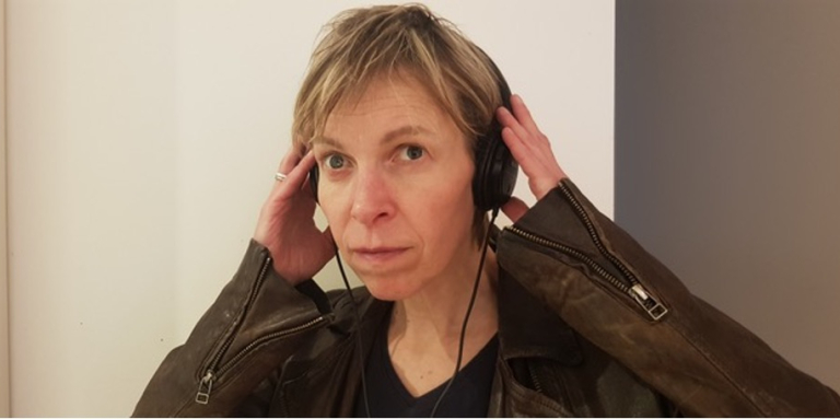 Photo en gros plan d'Alex regardant l'objectif. Elle pose ses deux mains sur le casque qu'elle porte. Elle porte une veste en cuir marron dotée de fermetures éclair aux poignets.