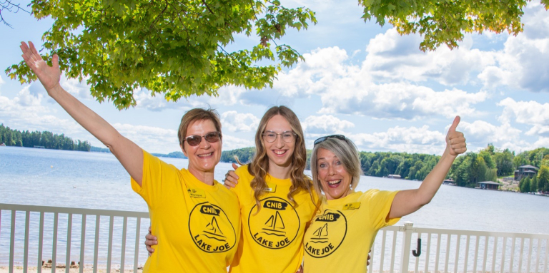 Monique Pilkington, Emma Van Dyk et Sherri Helsdingen se tiennent côte à côte au bord du lac Joe, vêtues de t-shirts jaunes « Lake Joe », les bras autour les unes des autres. Elles sourient à la caméra et lèvent le pouce.